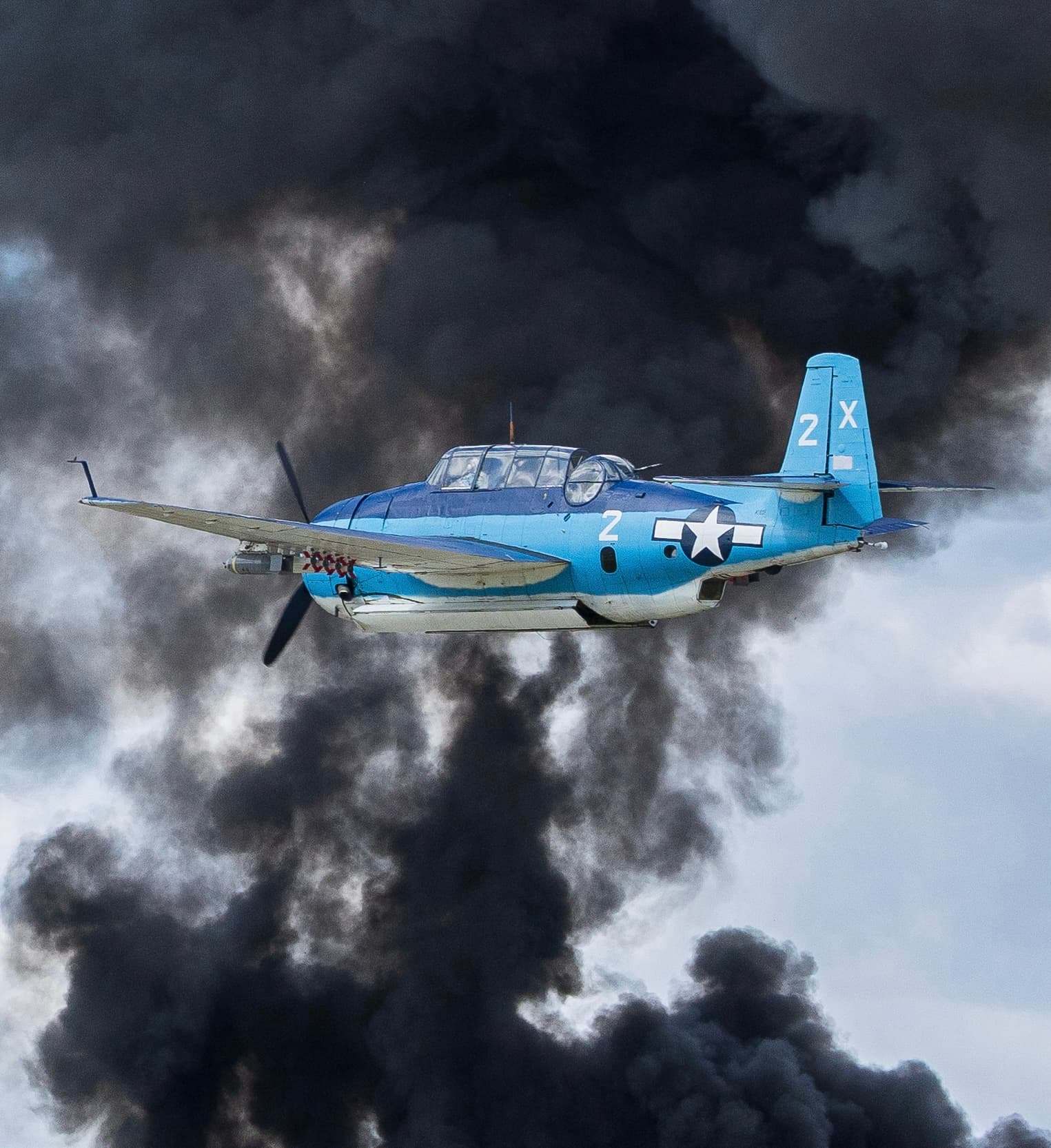 Blue WWII-era propeller aircraft flying through dark smoke clouds