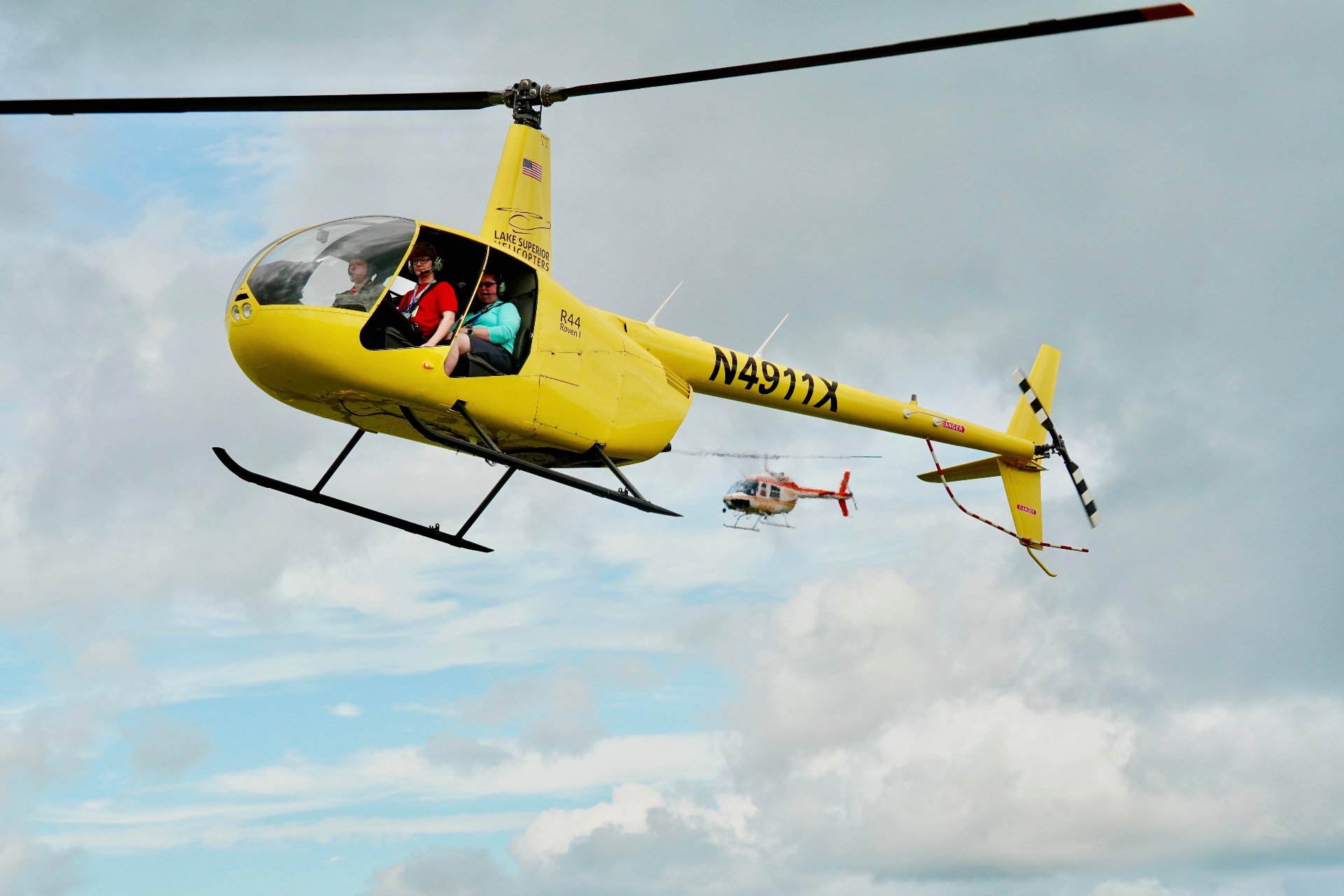 Yellow helicopter carrying passengers in flight, with a second helicopter visible in the cloudy sky behind it.