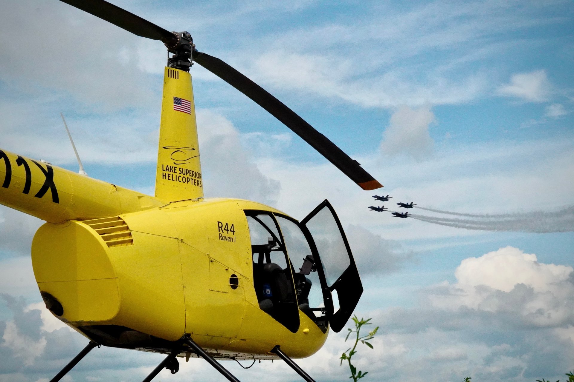 Yellow helicopter with its door open as a formation of jets flies past in the cloudy sky.