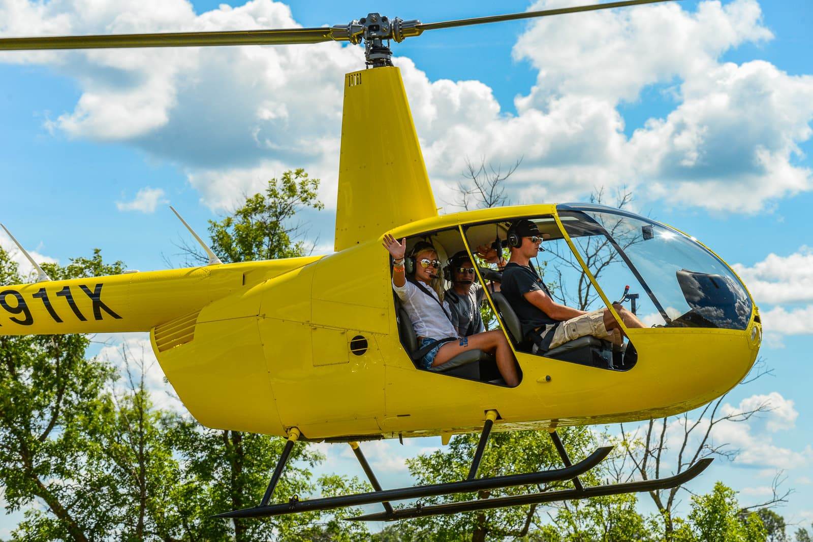 Passengers wave from a yellow helicopter during a ride beneath a bright, partly cloudy sky.
