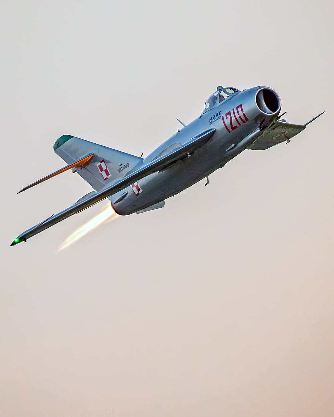 Silver MiG-17 jet climbing through the sky with afterburner lit, marked with red 1210 and Polish insignia.