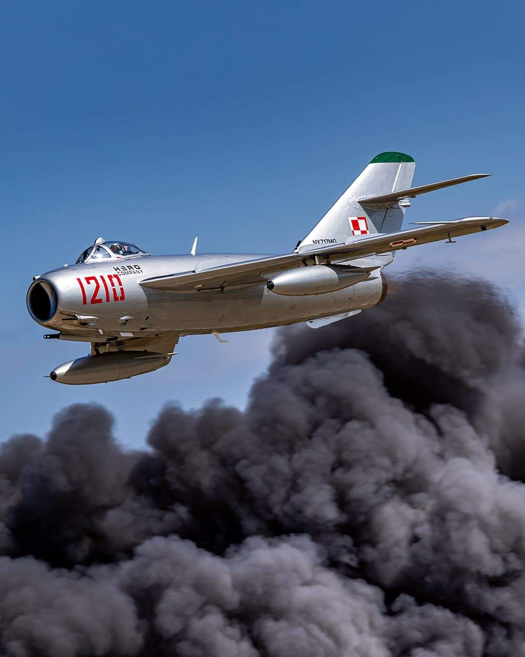 Silver MiG-17 jet in flight above a large plume of dark smoke, marked with red 1210 and Polish insignia.