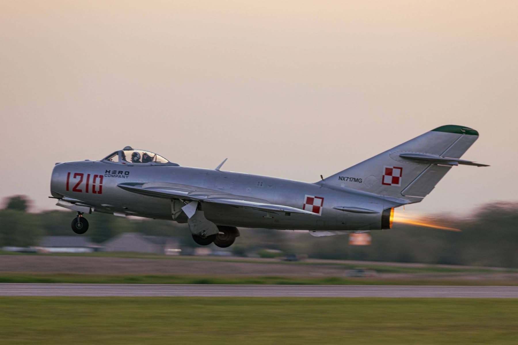 Silver MiG-17 jet flying low over a runway with landing gear down and afterburner glowing.