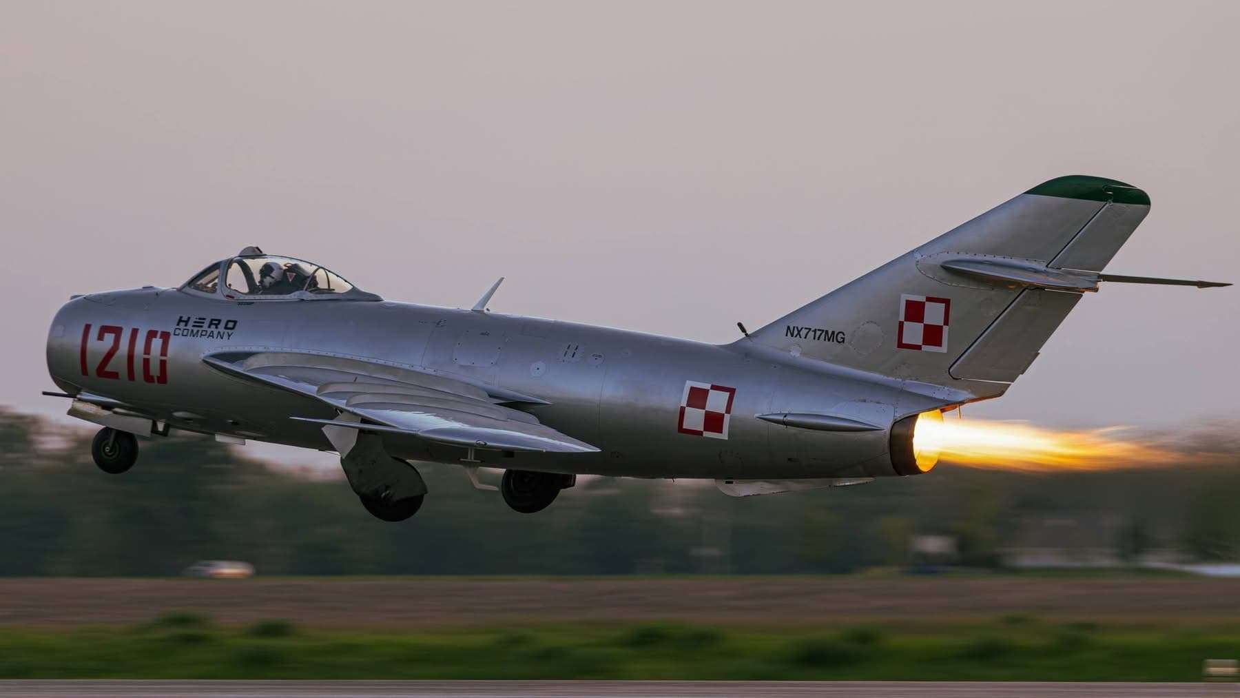 Side view of a silver MiG-17 jet flying just above the runway with landing gear down and afterburner lit.