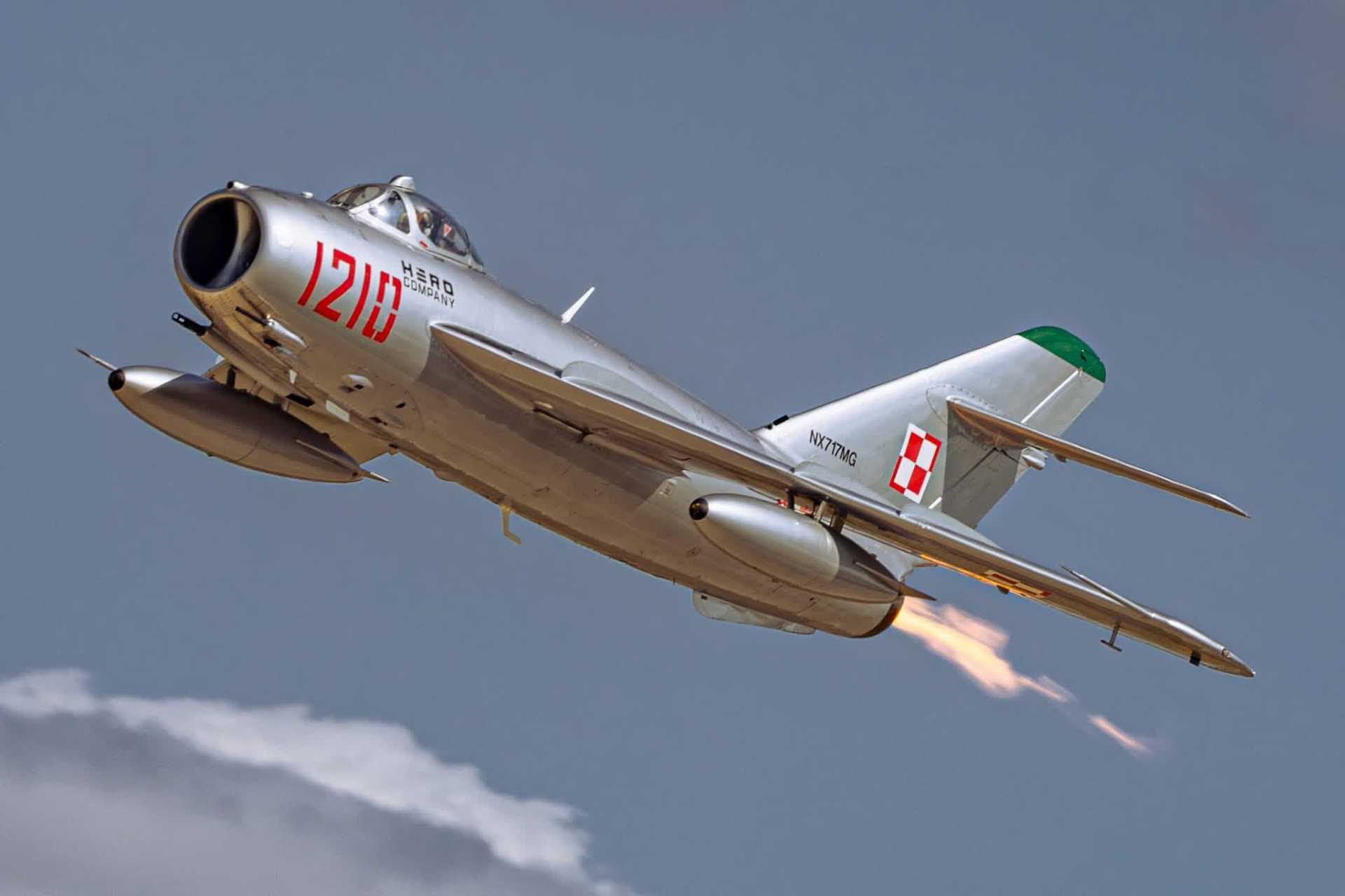 Silver MiG-17 jet climbing against a blue sky with afterburner lit, showing Polish insignia and red 1210 markings.