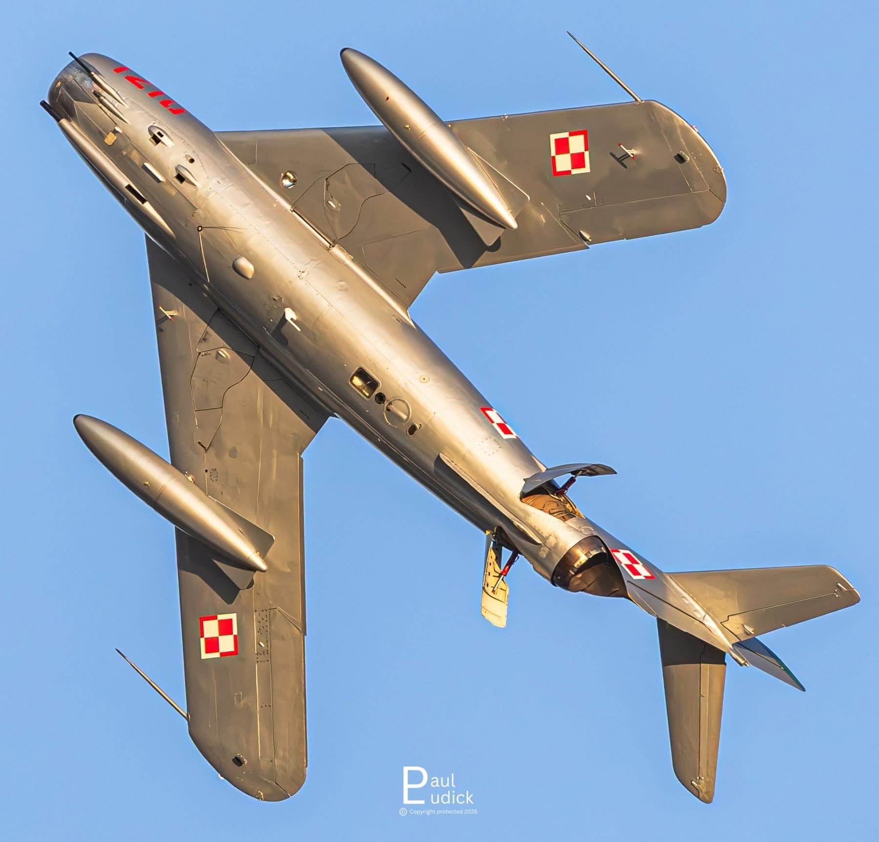 Underside view of a silver MiG-17 jet banking steeply against a clear blue sky, displaying red-and-white Polish insignia.