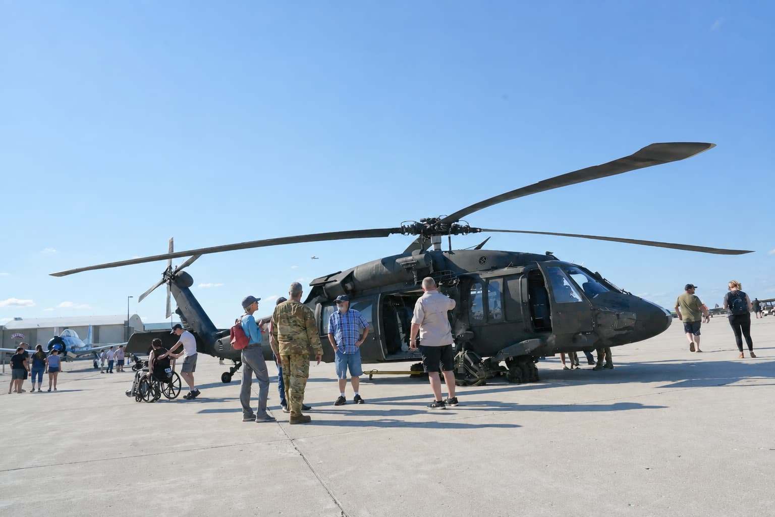Black military helicopter on display at an airshow, with visitors gathered around it on the tarmac under a clear blue sky.