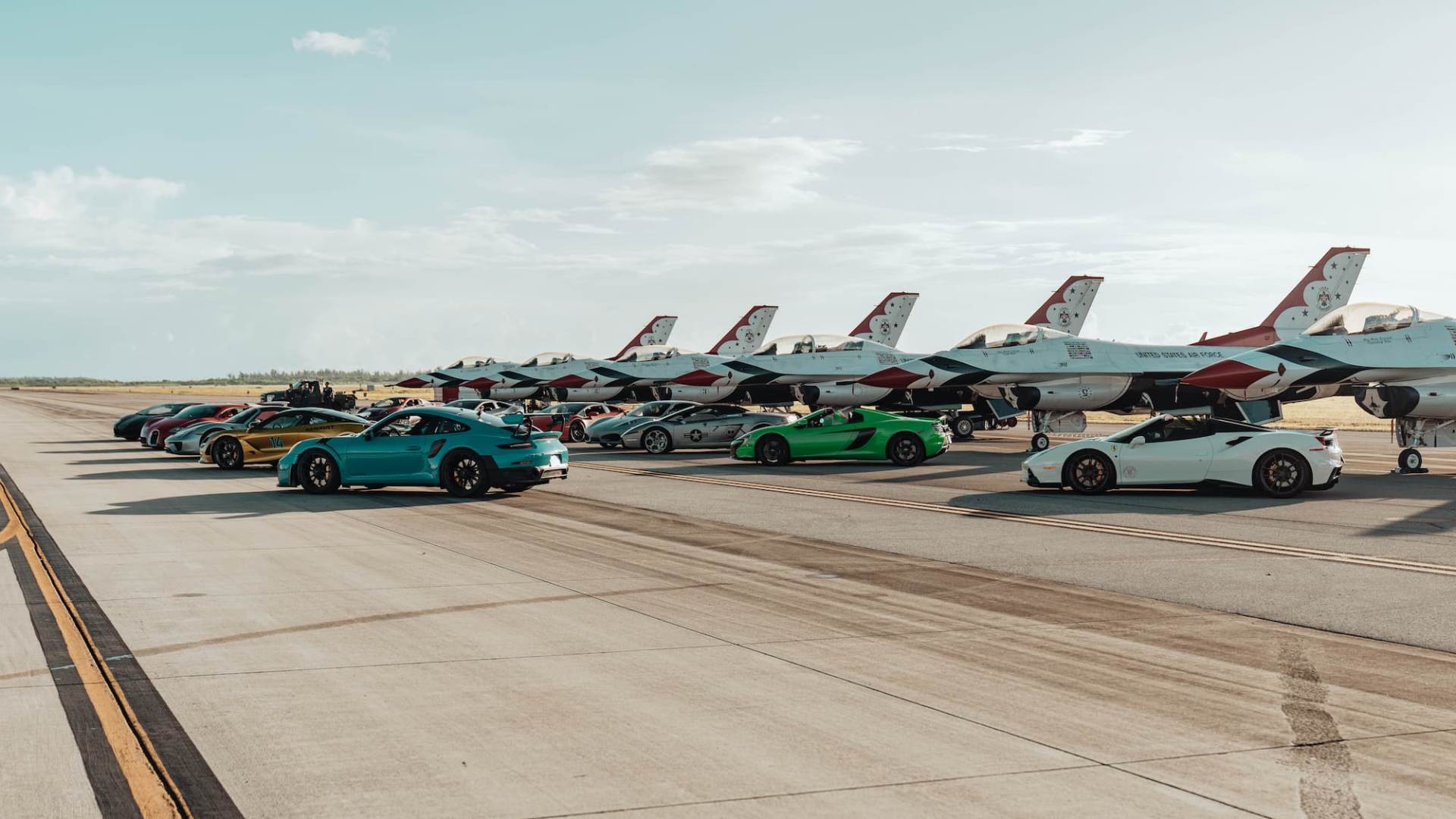 A row of colorful luxury sports cars is parked beside a lineup of U.S. Air Force Thunderbirds F-16 jets on an airstrip under a clear sky.