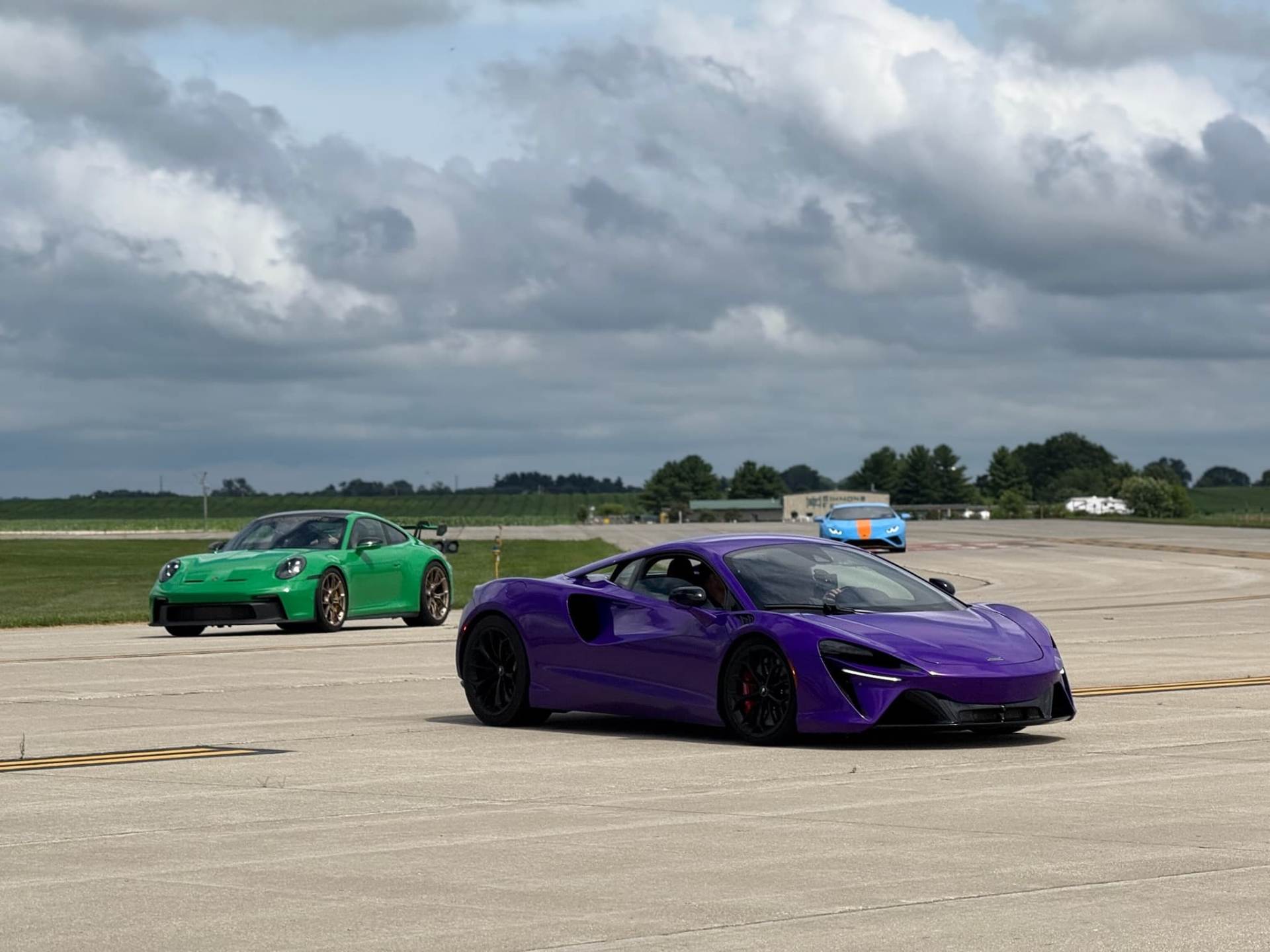 A line of brightly colored exotic cars parked on the runway in front of white event tents under a clear blue sky.