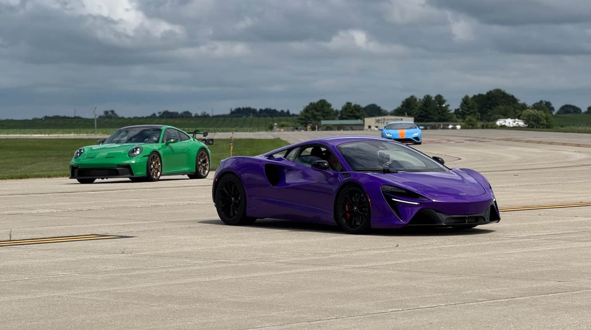 A line of brightly colored exotic cars parked on the runway in front of white event tents under a clear blue sky.