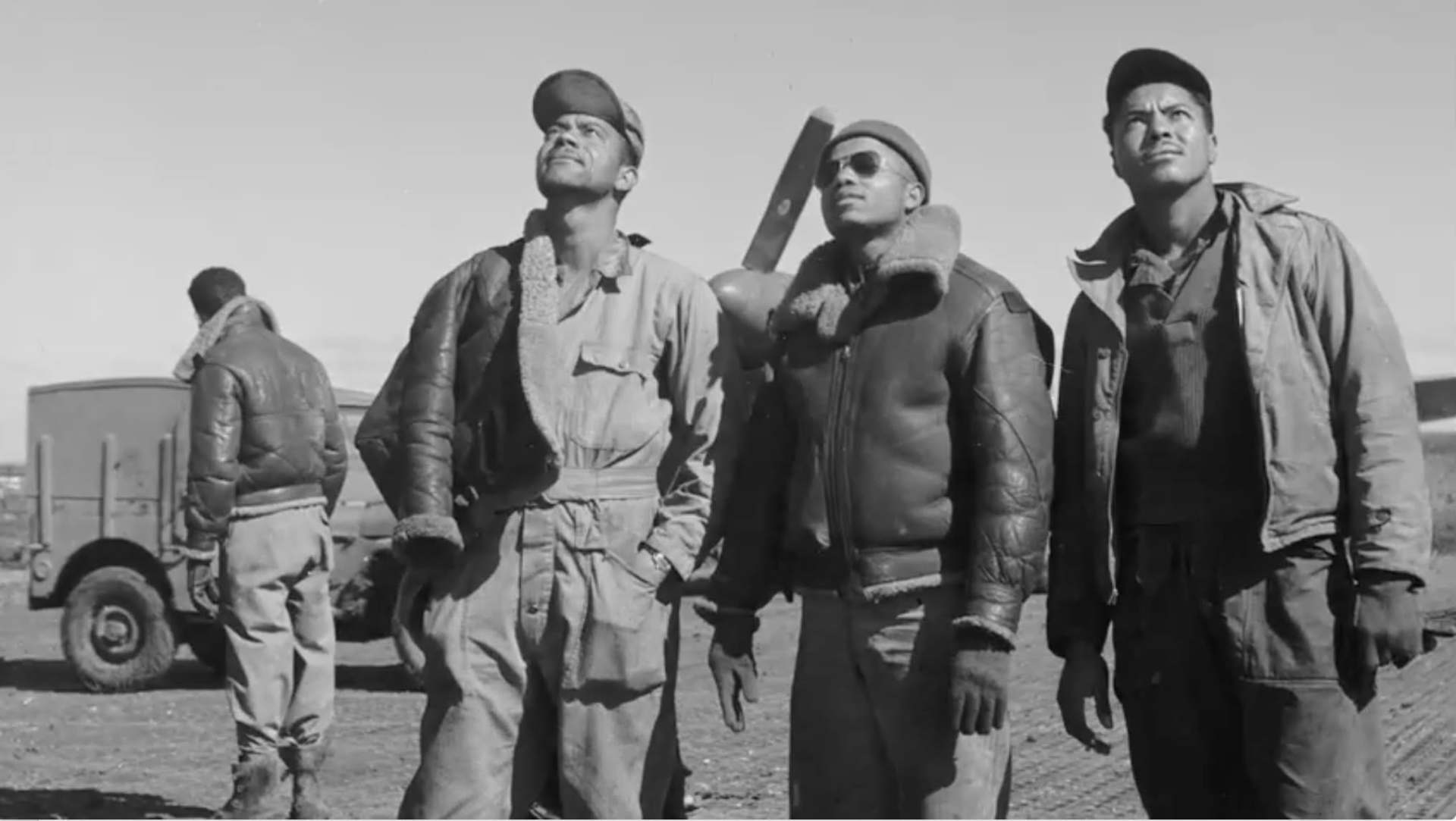 Three Tuskegee Airmen in flight gear looking upward beside an aircraft.