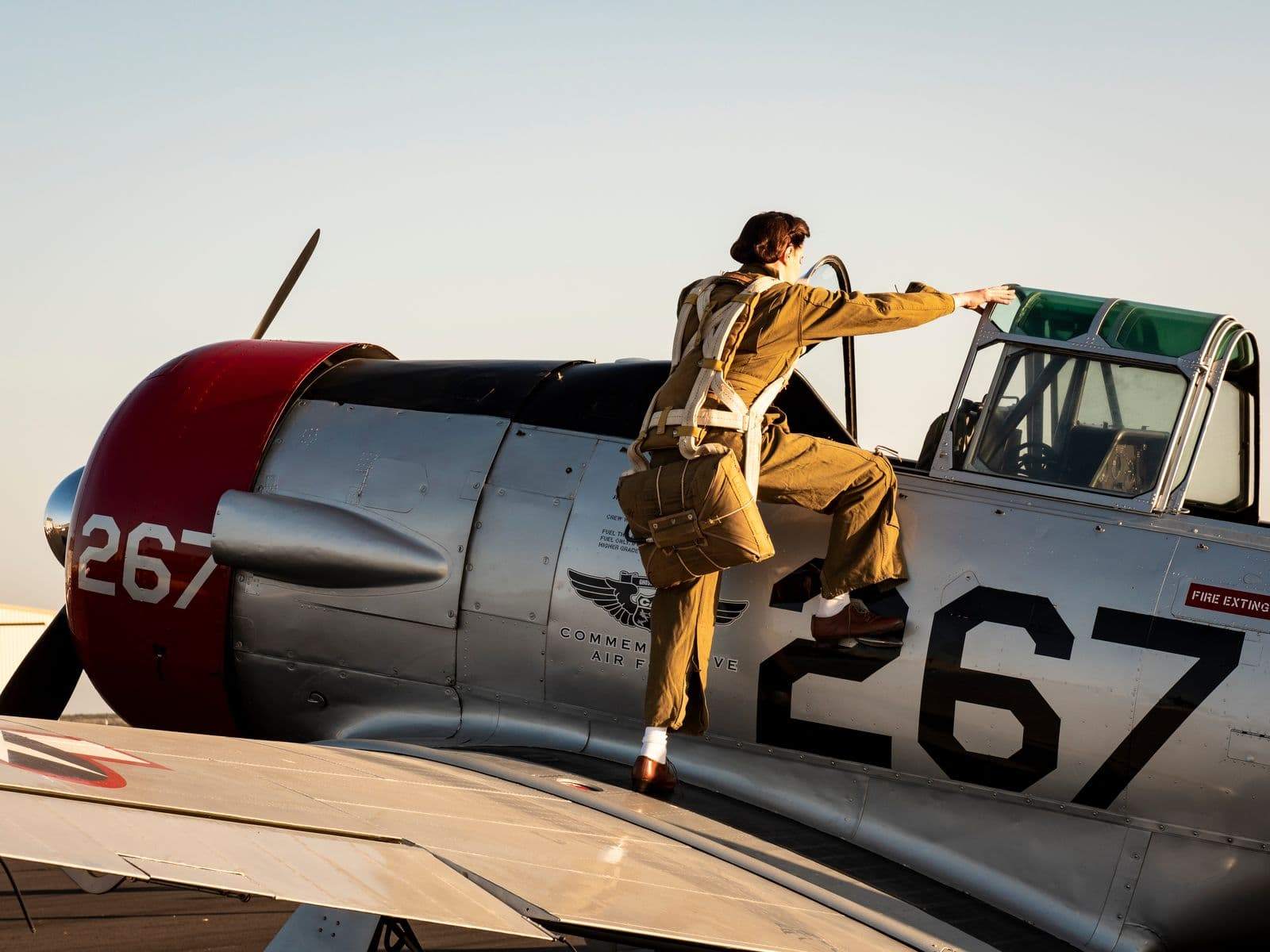 A pilot in World War II-style flight gear climbs onto the wing and reaches into the cockpit of a silver vintage aircraft numbered 267.