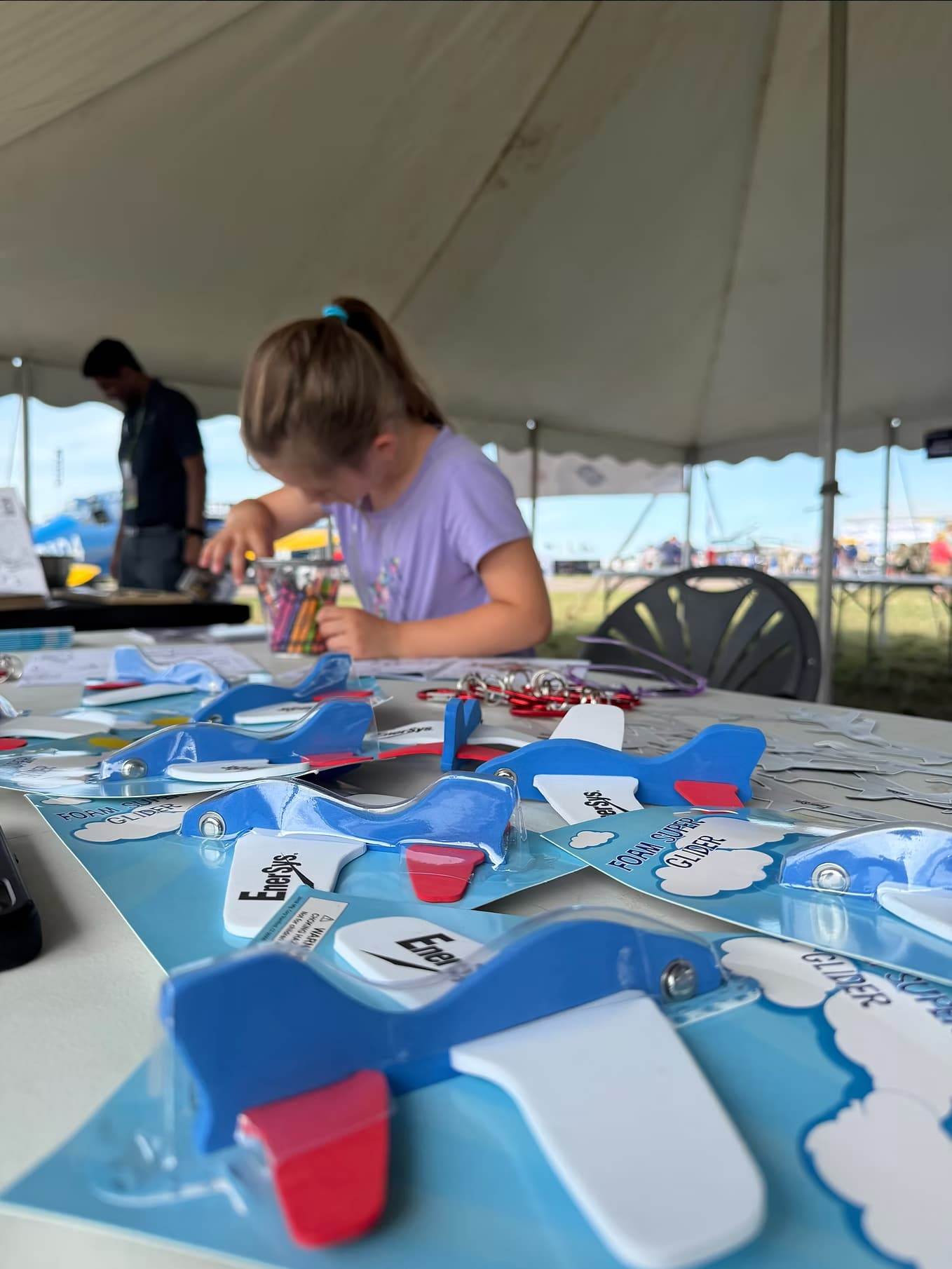 Foam airplane glider kits and craft materials on a table while a child assembles an aircraft model.