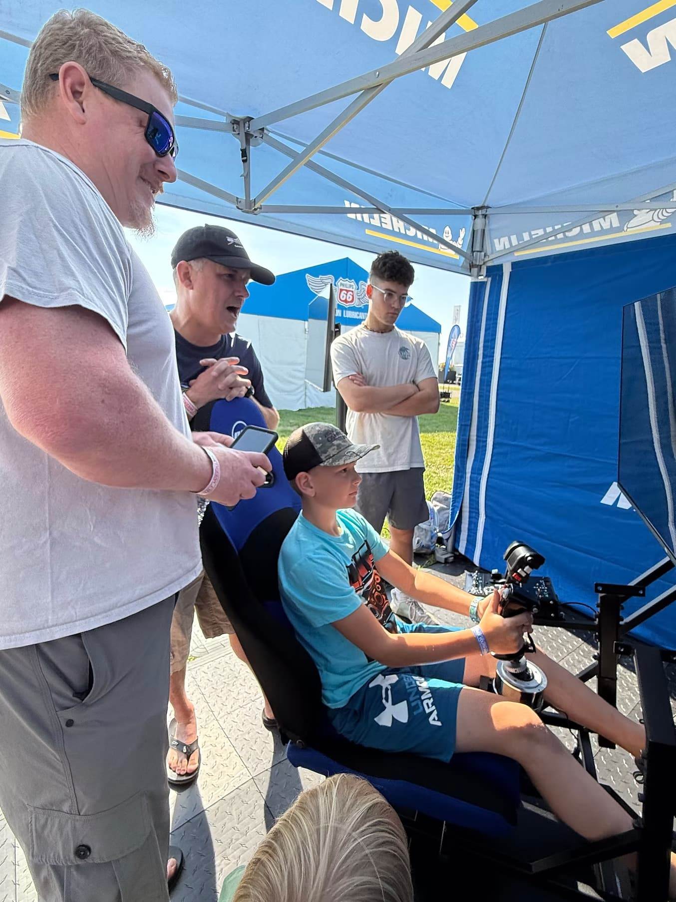 Young participant using a flight simulator while adults watch and assist inside an aviation exhibit tent.