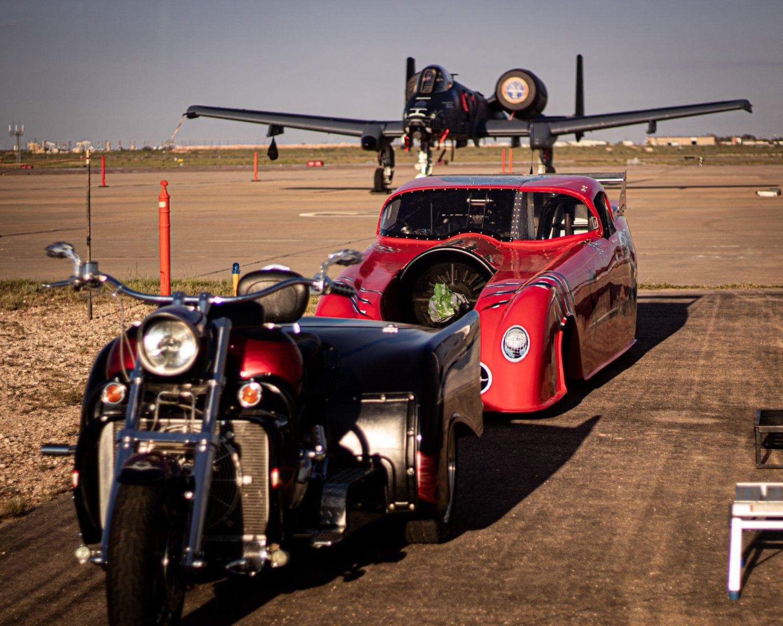 Slayer Jet Car parked behind a custom trike on a runway with a military aircraft in the background