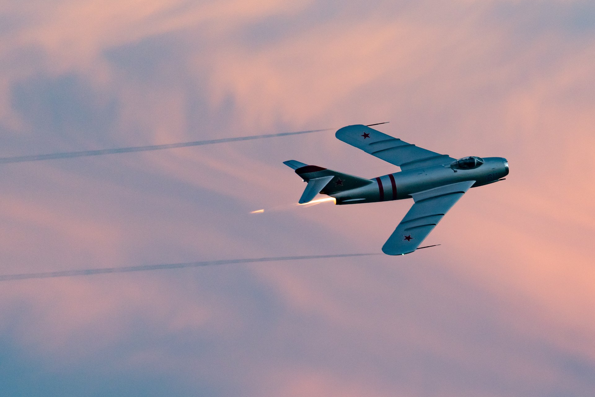 A MiG-17F jet performs a fast banking pass at sunset with its afterburner glowing and vapor trails streaming behind it.