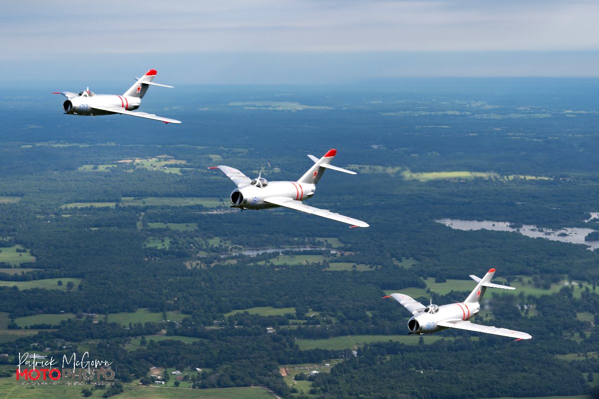 Three MiG-17F fighter jets flying in formation over a green, rural landscape.