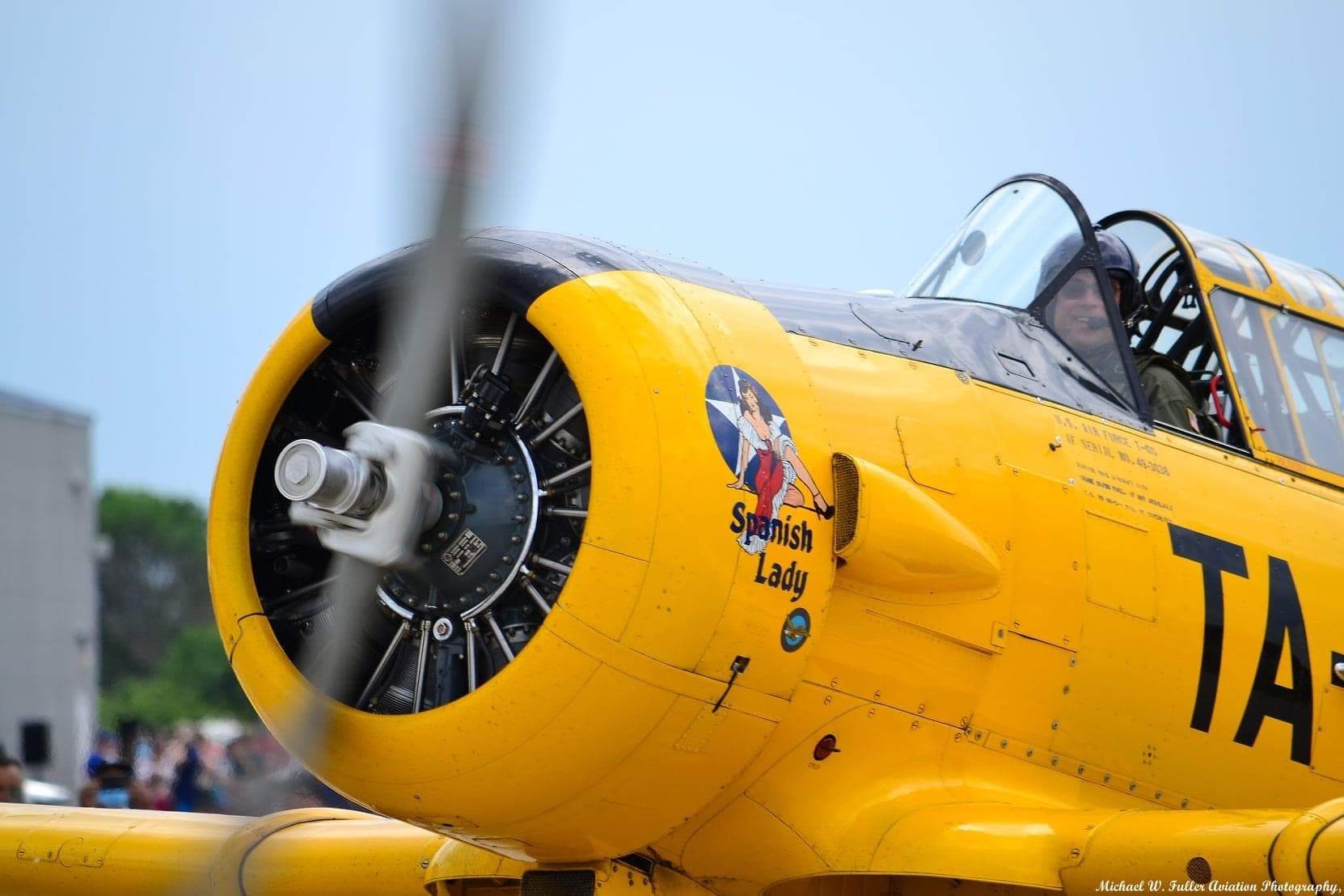 Close-up of the yellow T-6 Texan radial engine and nose with spinning propeller, showing the Spanish Lady artwork and pilot in the cockpit.