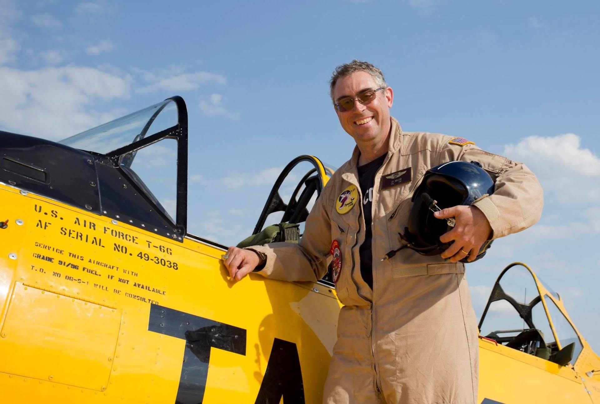 Pilot standing beside a yellow T-6 Texan aircraft, smiling and holding a helmet, with the open cockpit visible behind him.