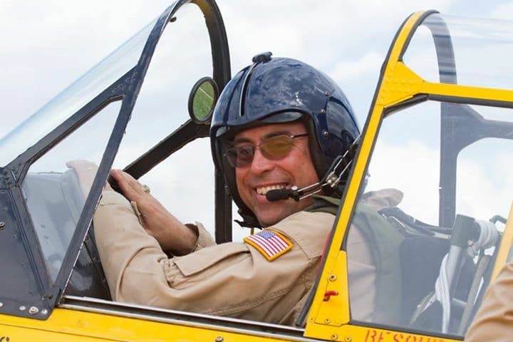 Pilot seated in the cockpit of a yellow T-6 Texan, smiling and wearing a flight helmet with headset.