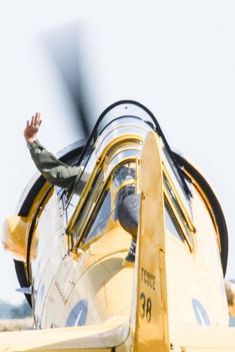 Front view of a yellow T-6 Texan with the pilot waving from the cockpit, propeller blurred in motion.