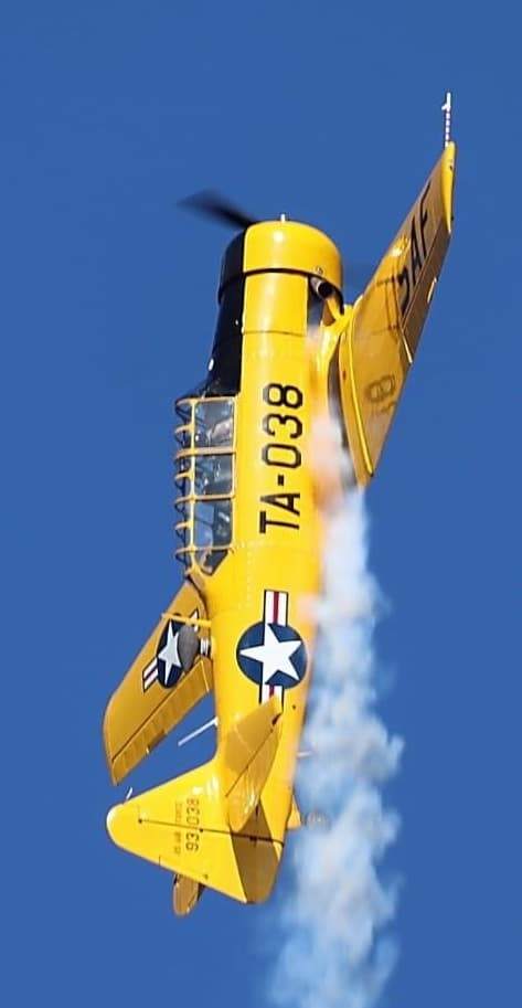 Yellow T-6 Texan in a steep vertical maneuver, trailing white smoke with U.S. Air Force markings visible against a clear blue sky.