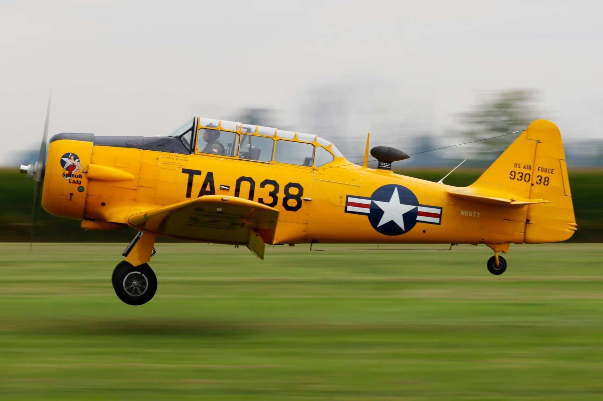 Side view of a yellow T-6 Texan aircraft in flight just above the runway, showing U.S. Air Force markings and motion blur in the background.