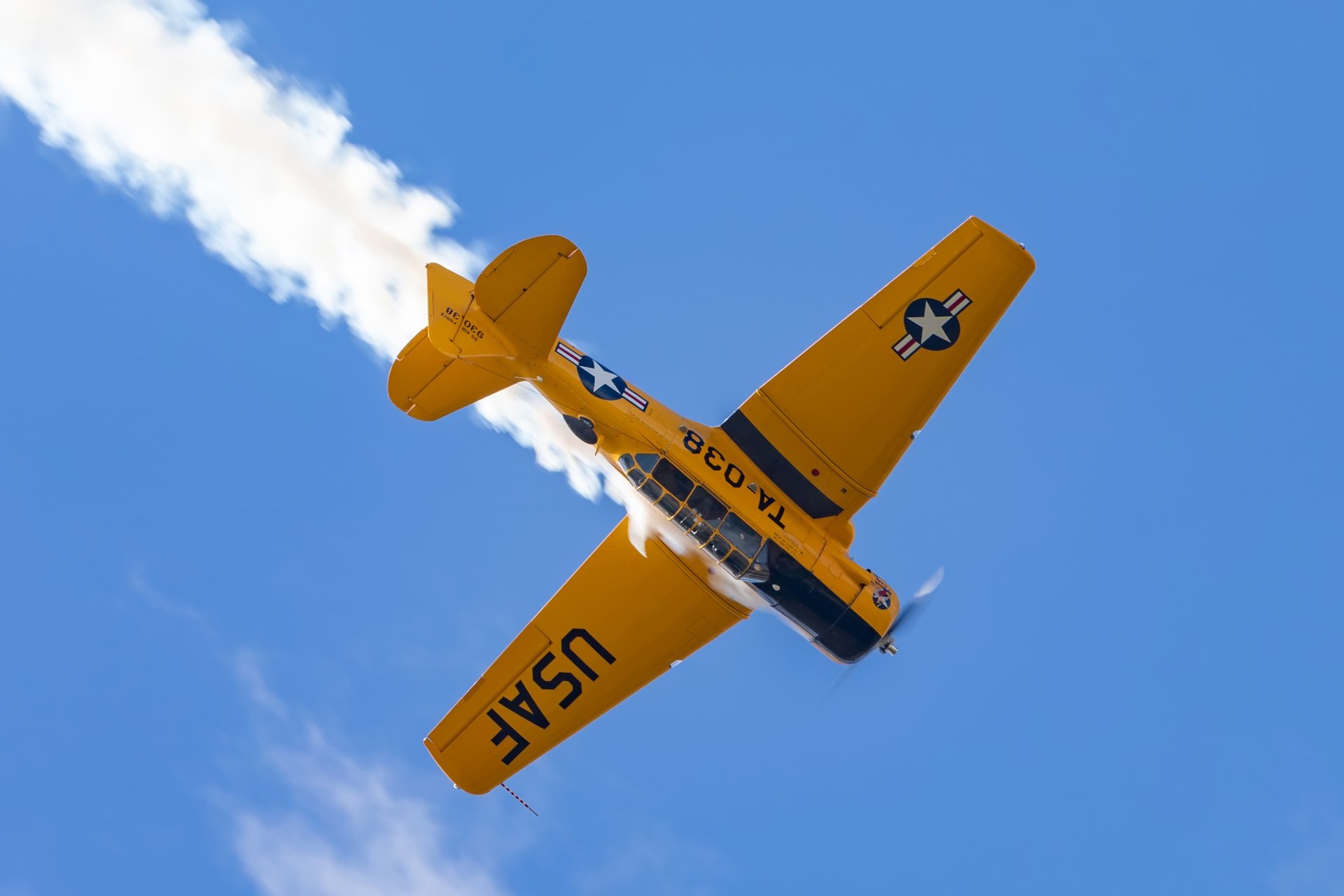 Underside view of a yellow T-6 Texan in flight, banking with white smoke trailing and USAF markings visible on the wings.