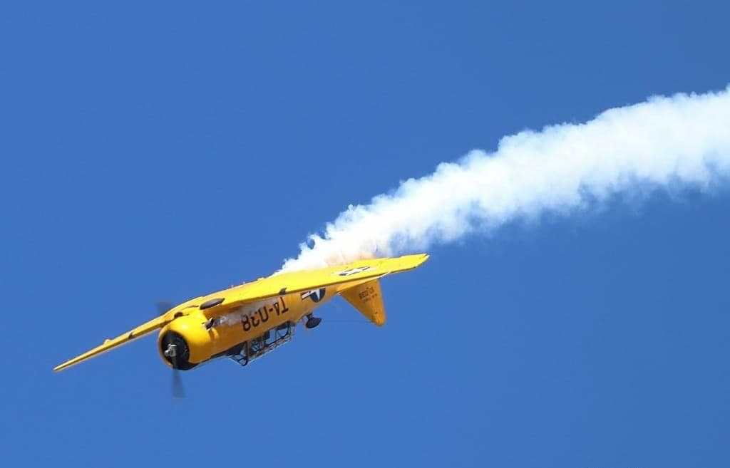 Yellow T-6 Texan aircraft performing an aerobatic maneuver in a clear blue sky, trailing white smoke behind it.