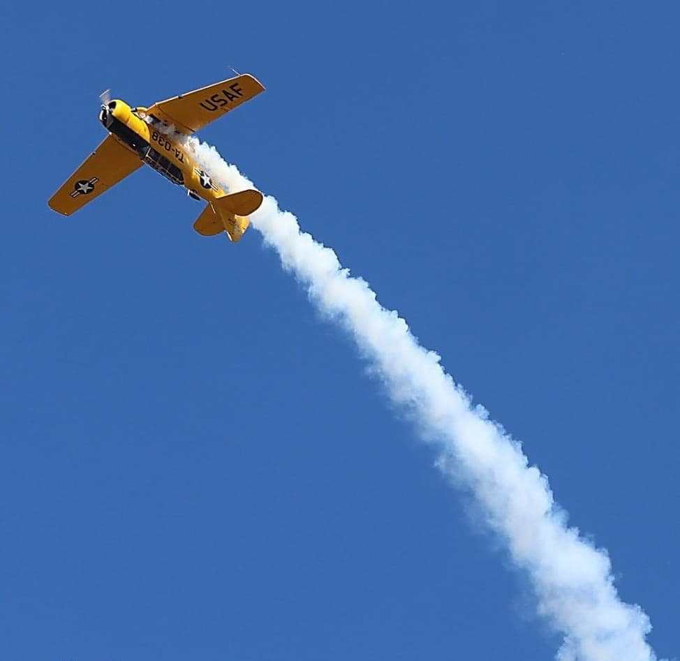 Yellow T-6 Texan climbing steeply during an aerobatic maneuver, leaving a long white smoke trail against a clear blue sky.