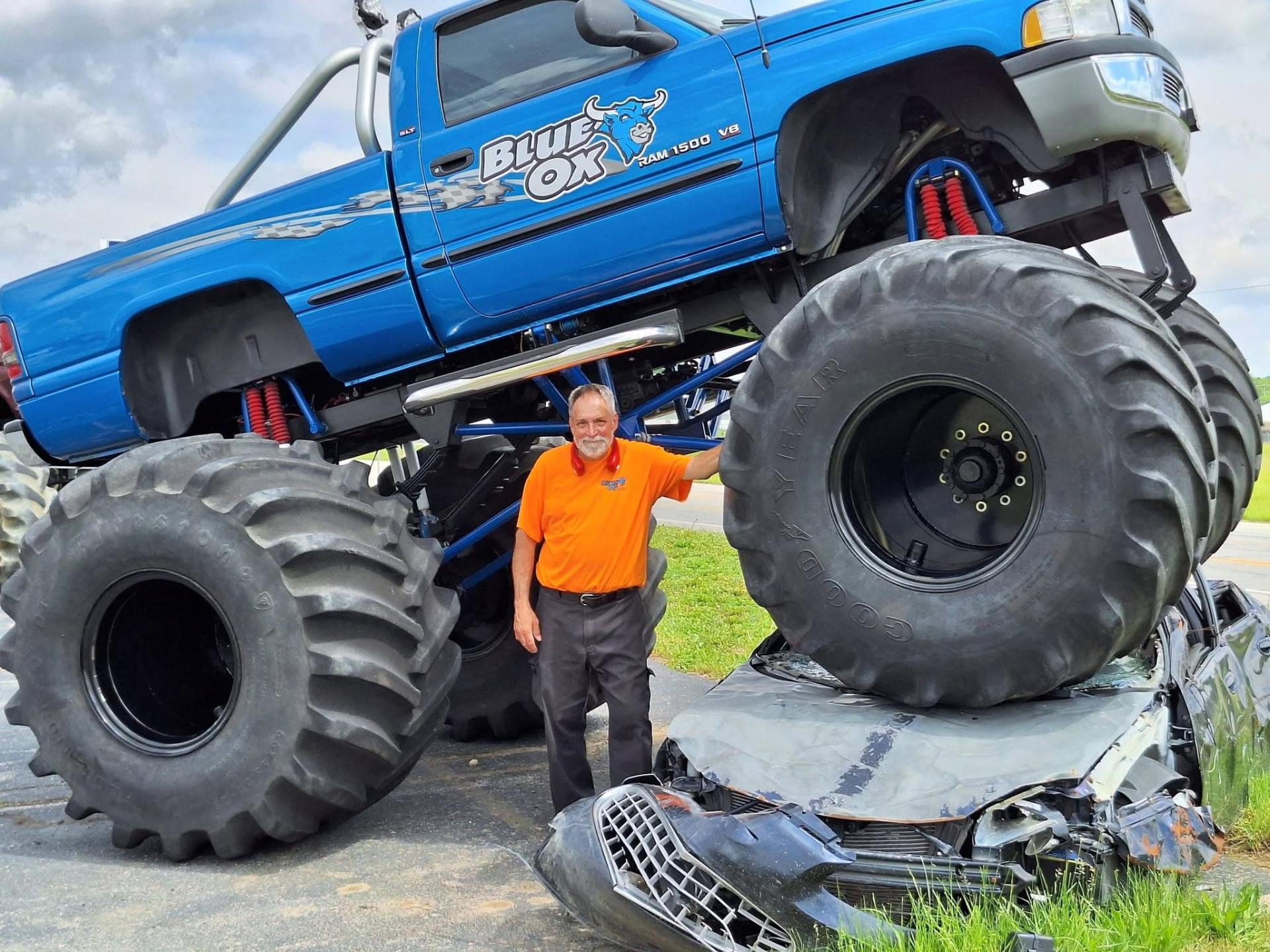 Blue Ox monster truck crushing a car, with a man standing beside one of its giant tires.