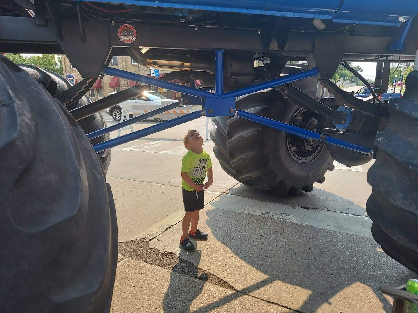 Young child standing beneath a towering blue monster truck, looking up at its suspension and enormous tires.