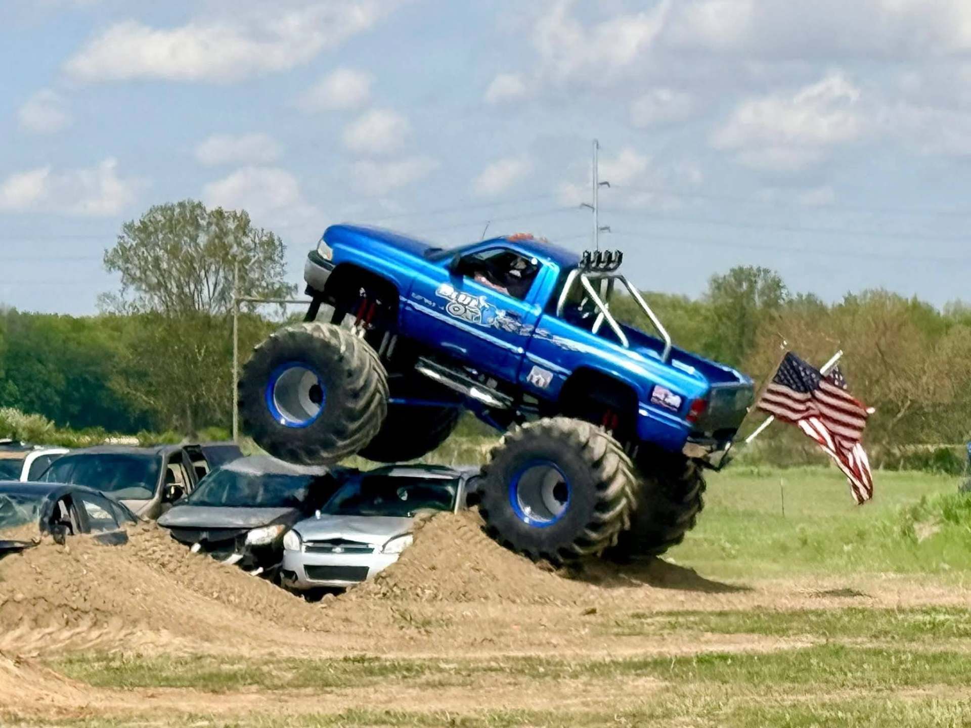 Blue monster truck driving over crushed cars, with two American flags flying from the rear.