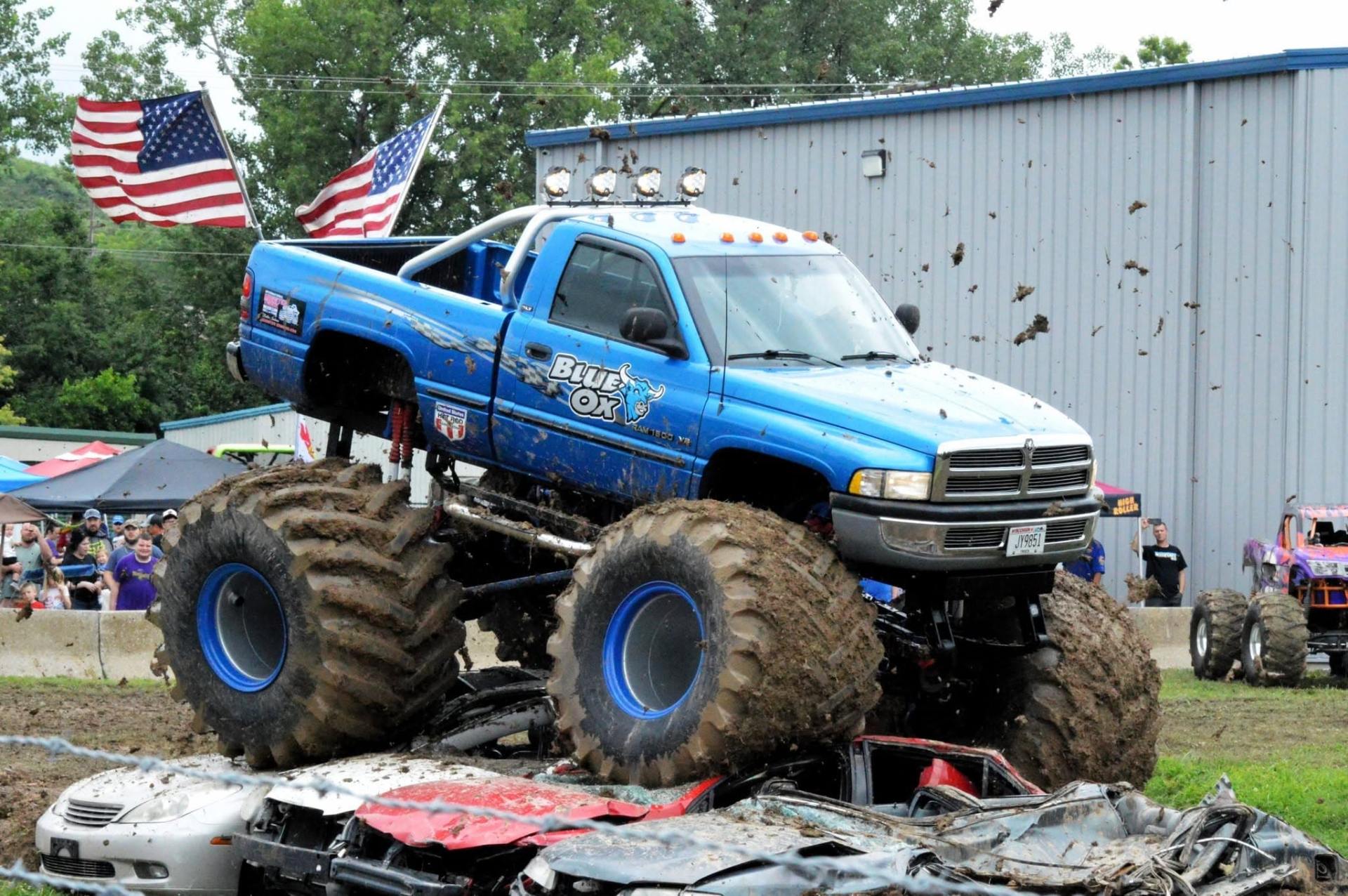 Blue Ox monster truck driving over crushed cars in a muddy arena, with two American flags flying from the truck.