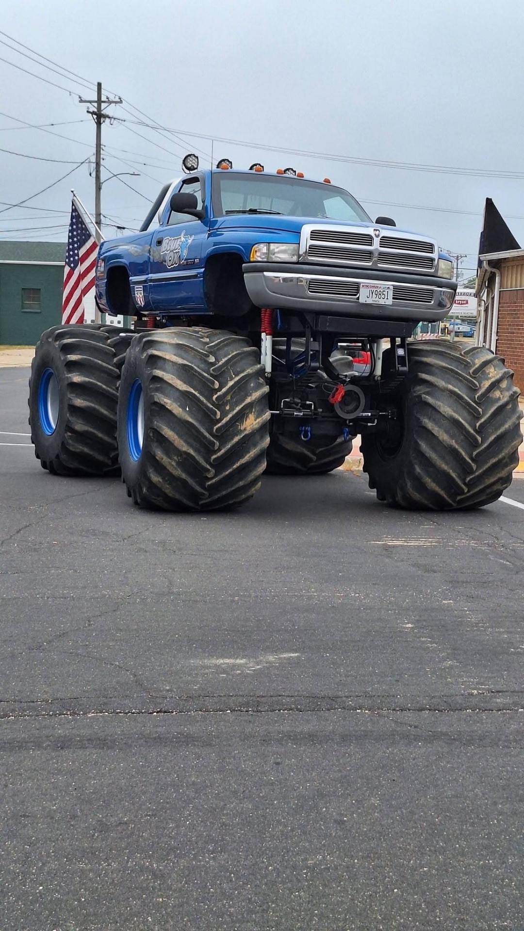 Blue Bear Foot monster truck parked on a street, with oversized tires and an American flag mounted at the rear.