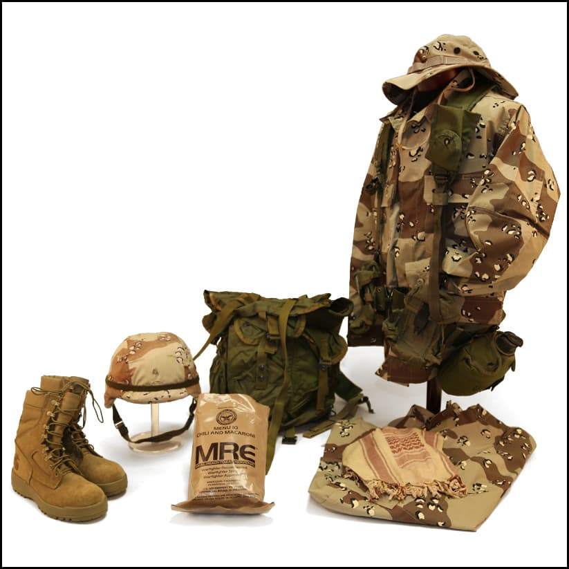 Desert Storm military field gear displayed with camouflage jacket, helmet, boots, pack, MRE, scarf, and folded uniform pieces.