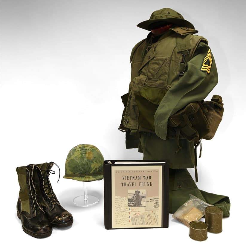 Vietnam War military field gear displayed with green uniform, helmet, boots, pack, food tins, and a travel trunk binder.