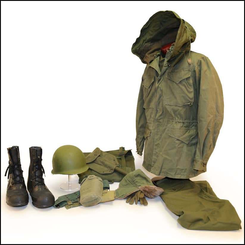 Military cold-weather field gear displayed with green parka, helmet, boots, gloves, canteen, and folded uniform pieces.