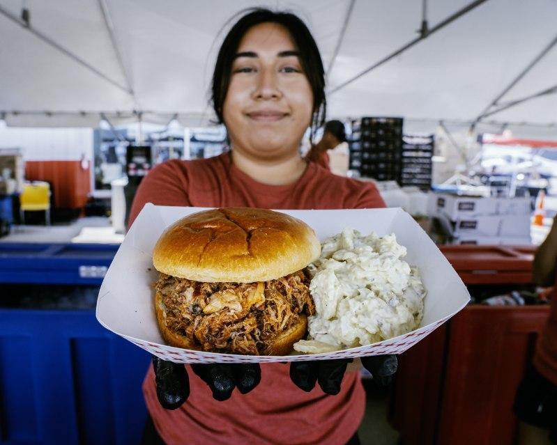 A food vendor holds a tray with a pulled pork sandwich and coleslaw at an outdoor event