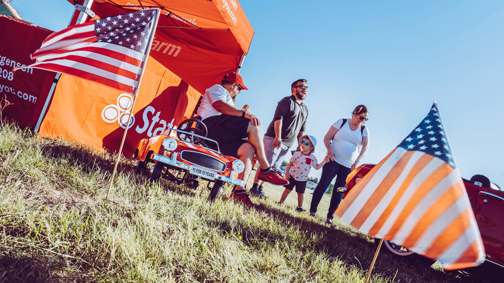 A family strolls through sponsor displays and vintage cars beneath American flags at an outdoor airshow