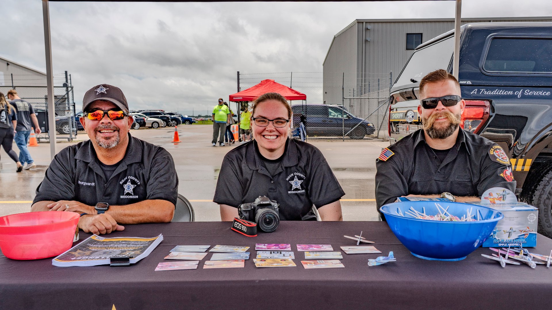 Three uniformed public safety staff sit behind an information table with giveaways at an outdoor airshow
