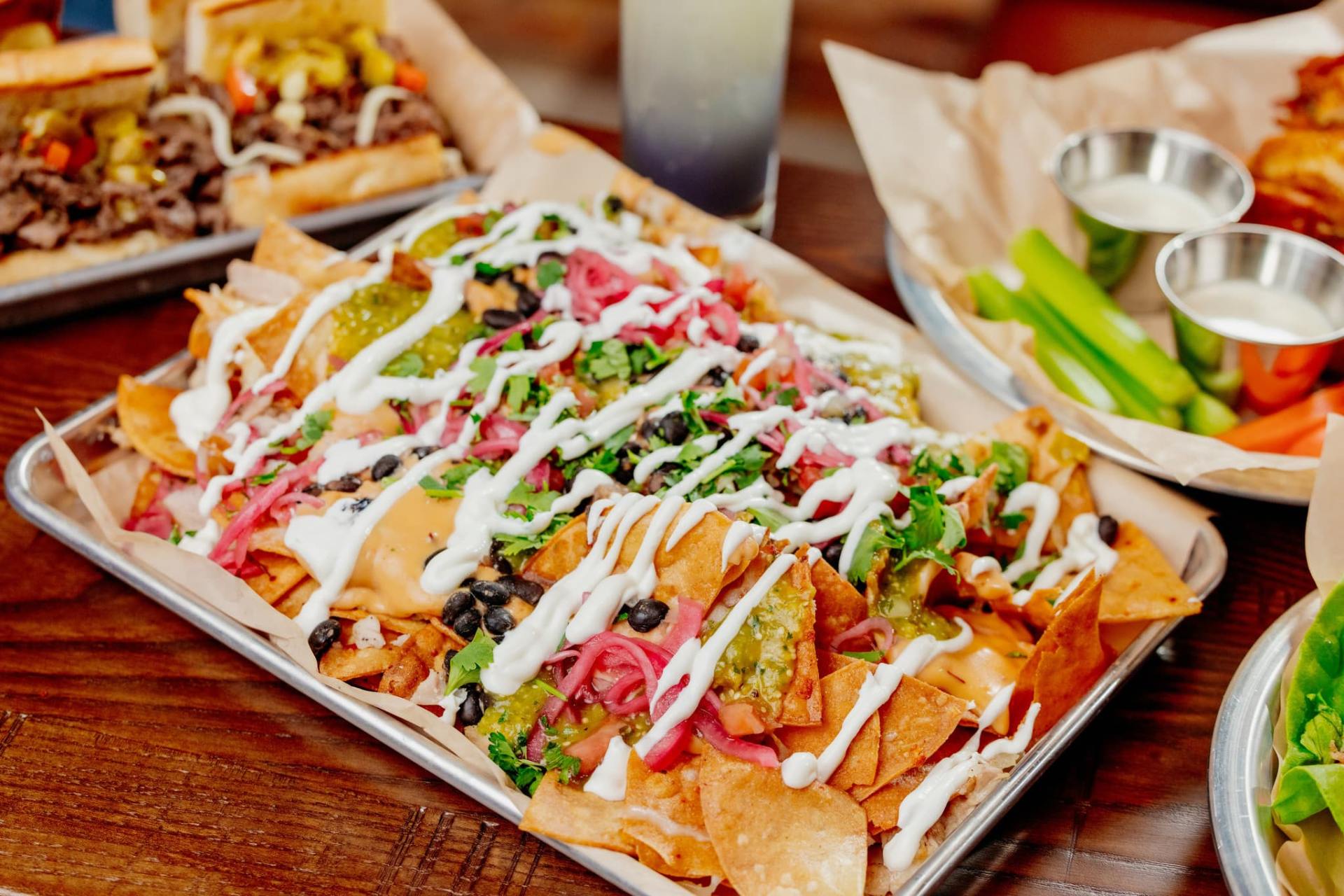 Tray of loaded nachos topped with cheese sauce, black beans, salsa verde, pickled onions, cilantro, and crema, with celery and dipping sauce in the background.