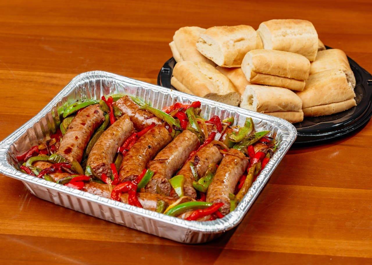 Tray of cooked sausages with peppers and onions, served with sliced bread rolls on a platter.