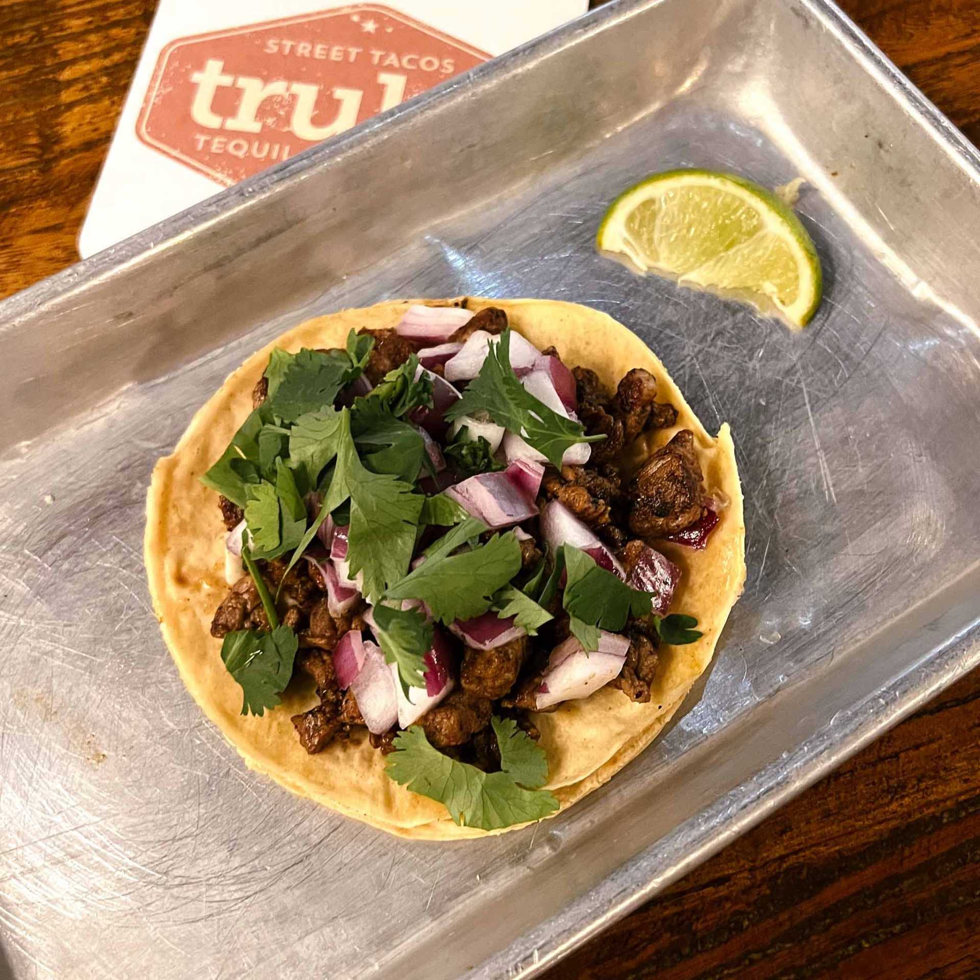 Street taco with chopped meat, red onion, and cilantro on a corn tortilla, served on a metal tray with a lime wedge.
